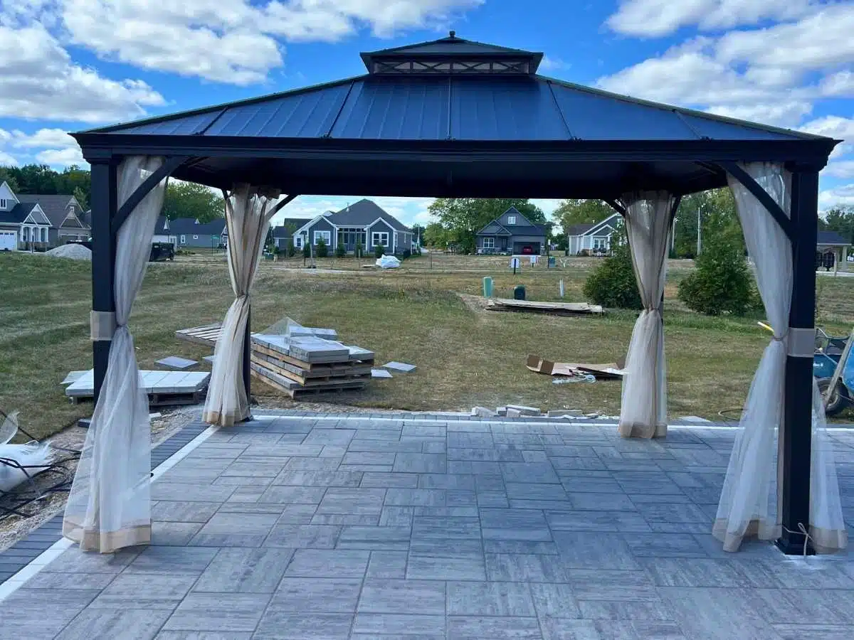 Black metal gazebo with light curtains installed on a large tiled patio in a residential backyard