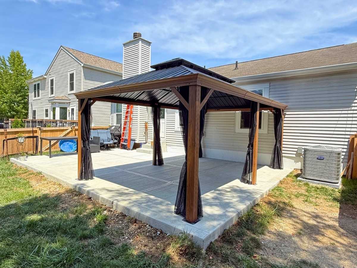 Metal roof gazebo with wood tone frame and curtains installed over a backyard concrete patio