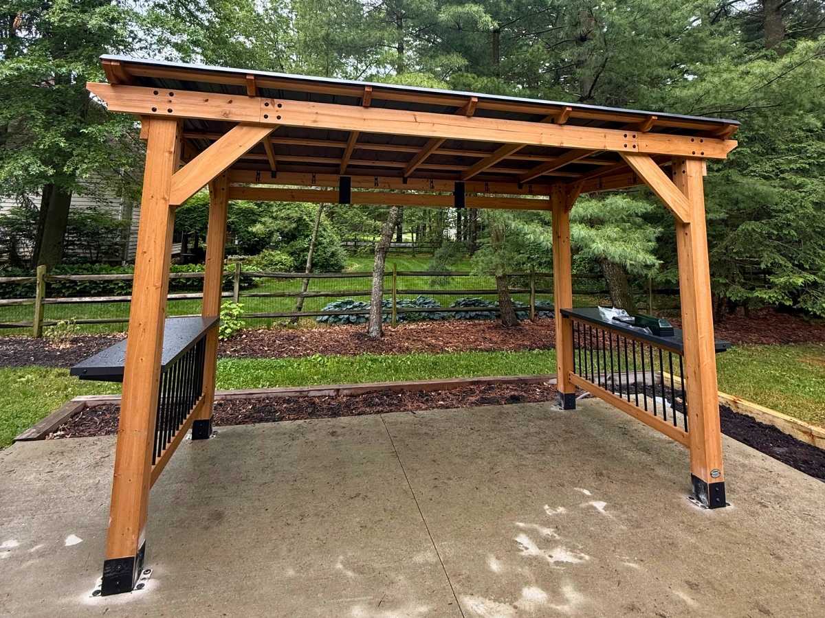 Wood pergola style gazebo with open roof design installed over a concrete patio surrounded by trees