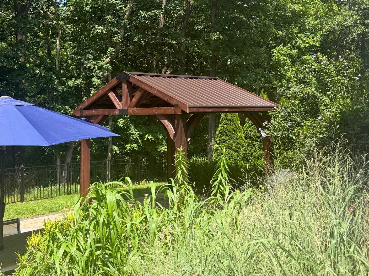 Finished wood pavilion style gazebo with metal roof in a landscaped backyard surrounded by trees